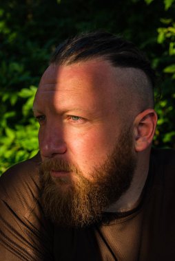 Yüzünde ciddi bir ifadeyle ağaca yaslanan bir adam profili. Ağaçta sakallı bir adamın profili. Ormanda sakallı yakışıklı bir adamın portresi.