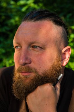 Yüzünde ciddi bir ifadeyle ağaca yaslanan bir adam profili. Ağaçta sakallı bir adamın profili. Ormanda sakallı yakışıklı bir adamın portresi.