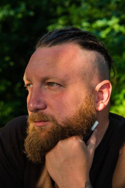 Yüzünde ciddi bir ifadeyle ağaca yaslanan bir adam profili. Ağaçta sakallı bir adamın profili. Ormanda sakallı yakışıklı bir adamın portresi.