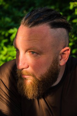 Yüzünde ciddi bir ifadeyle ağaca yaslanan bir adam profili. Ağaçta sakallı bir adamın profili. Ormanda sakallı yakışıklı bir adamın portresi.