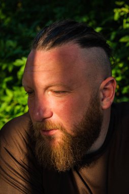 Yüzünde ciddi bir ifadeyle ağaca yaslanan bir adam profili. Ağaçta sakallı bir adamın profili. Ormanda sakallı yakışıklı bir adamın portresi.