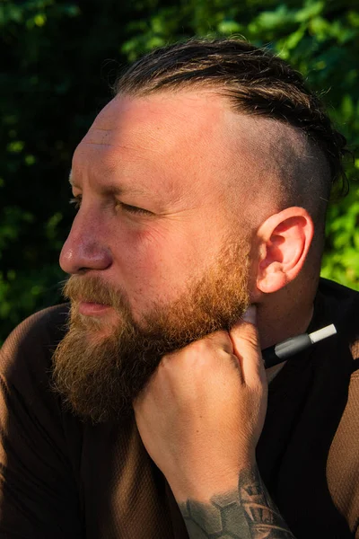 Profile view of man leaning against tree with serious expression on face. Profile of a man with a beard on a tree background. Portrait of handsome bearded man in forest