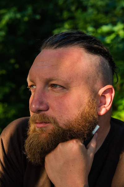 Yüzünde ciddi bir ifadeyle ağaca yaslanan bir adam profili. Ağaçta sakallı bir adamın profili. Ormanda sakallı yakışıklı bir adamın portresi.