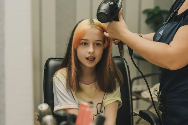Hairdresser making a hair style to cute little girl. A teenage girl has her bangs lightened in a beauty salon. Orange bangs. The trend of bright hair.