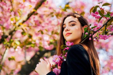 Uzun saçlı kız, çiçek açan sakura ağacının yanında bahar doğasının güzelliğinin tadını çıkarıyor..