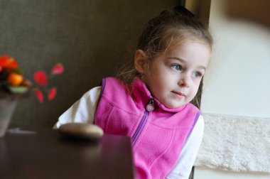 Bir kafede pencerenin yanında oturan küçük kız. Pencereden gün ışığında bir çocuk masada oturuyor.