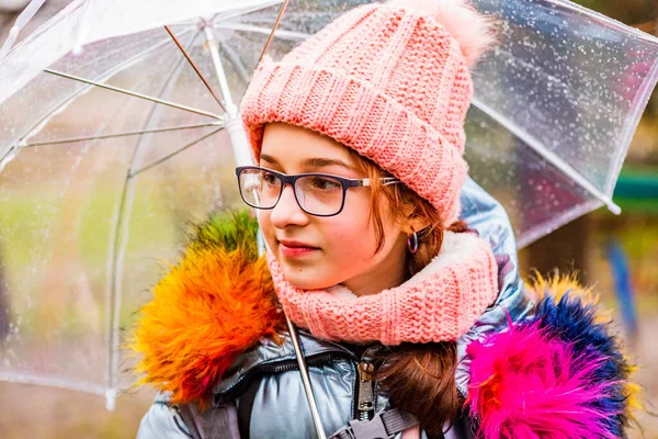 girl in a jacket with a transparent umbrella in the rain in the cold. Teenager under an umbrella