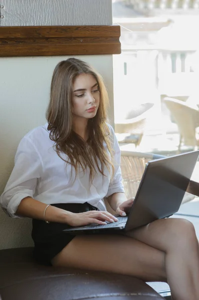 woman working behind laptop near window in cozy atmosphere. Girl in a white shirt and black shorts.