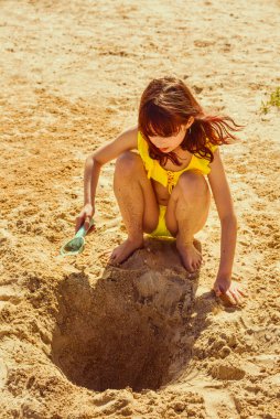 Sarı mayolu küçük bir okul kızı deniz kenarında kumdan bir şey inşa ediyor..