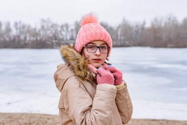 Pembe şapkalı ve paltolu bir kızın portresi kış manzarasında duruyor. Gözlüklü kız. nehir