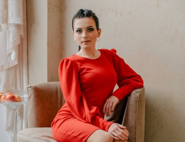 Young girl in a red dress indoors. model. Emotions of a modern lady. The girl is sitting in a chair