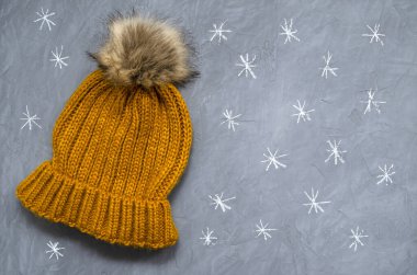 Gri beton arka plan manzaralı hardal renkli kürk ponponlu küçük şirin örülmüş, sıcak yün bir şapka. Tebeşirle çizilmiş küçük kar taneleri. metin için yer ile