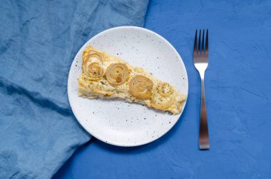 Aperatif balık fileto dilimi soğan ve otlarla pişirilmiş bir tabakta, peçete ve çatalla mavi bir zemin üzerinde.