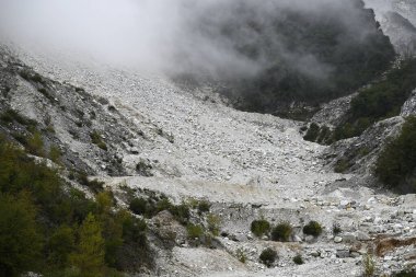 Apennine Dağları 'ndaki Carrara Mermer Madeni. Biri FG 'yi, işçileri, tekerlek yükleyicisini, mermer detaylarını ve bir köprüyü görüyor. Büyük bir taşın üzerinde koruyucu kasklar var..