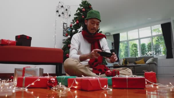微笑的亚洲男人 用手机拿着一个照片礼品盒 在家里的客厅里上传到网上 背景上有一棵圣诞树 圣诞节庆祝概念 — 图库视频影像