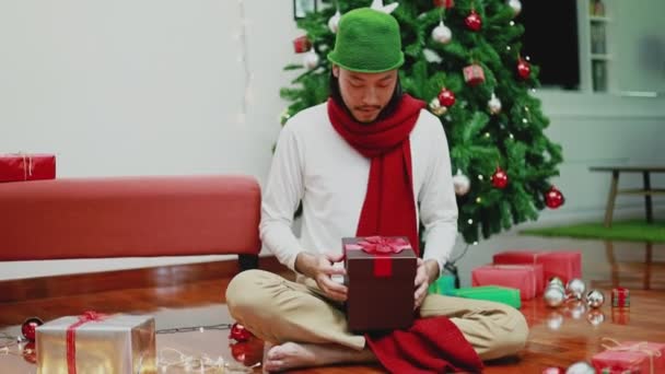 令人惊讶的是 亚洲男人打开了家中客厅里的礼品盒 后面有一棵圣诞树 圣诞节庆祝概念 — 图库视频影像
