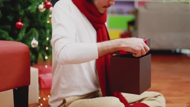 在家里客厅里 年轻的亚洲男人打开礼品盒 背后有一棵圣诞树 圣诞庆祝概念 — 图库视频影像