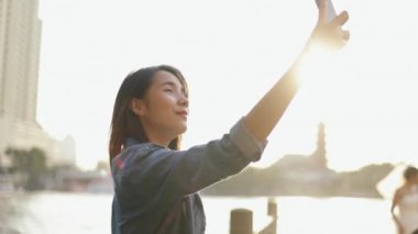 Güzel Asyalı kadın arkadaşlarıyla görüntülü konuşma yapıyor yaz tatilini paylaşarak Tayland 'da Chao Phraya Nehri' nin yanında durarak Bangkok Tayland 'da seyahat ediyor..