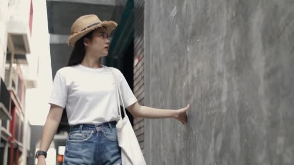 一位美丽的亚洲女人戴着一顶老式的帽子 提着一个布袋 用她的手轻柔地触摸着墙背 环视着一条小街道 享受暑假 — 图库视频影像
