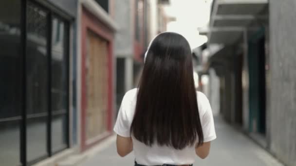 美丽的亚洲女人戴着耳机 一边听音乐一边跳舞 庆祝暑假 她走在老城区的一条小街上 — 图库视频影像