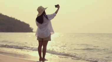 Sahilde gün batımında beklerken akıllı telefon kullanan Asyalı güzel genç bir kadın. Yaşam tarzı seyahat tatil yaz tatili konsepti.