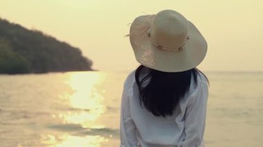 Fötr şapka takan çekici Asyalı kadın sahilde durmuş deniz manzaralı güzel gün batımını seyrediyor. Yaşam tarzı seyahat tatil yaz tatili konsepti.