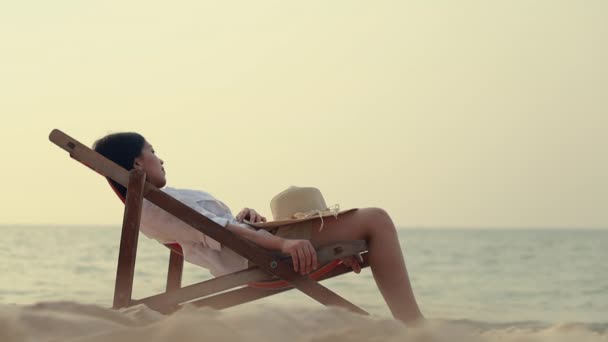 年轻的亚洲女人躺在沙滩上的躺椅上 感到很放松 她的脸紧闭着一顶夏威夷帽 暑假的概念 — 图库视频影像