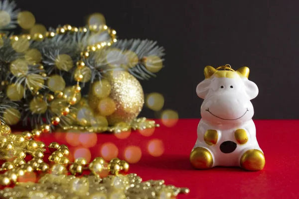 Estatuilla de una vaca rodeada de decoraciones navideñas y un árbol de Navidad sobre un fondo rojo y negro, tarjeta de Navidad 2021 año del toro — Foto de Stock