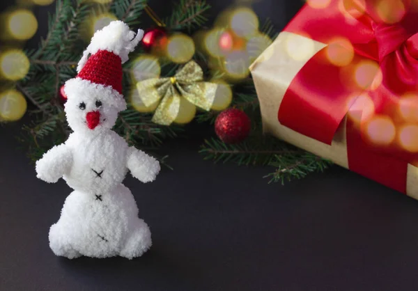 Fabulosa tarjeta de Año Nuevo sobre un fondo oscuro con un muñeco de nieve en un sombrero de Santa Claus rojo, bokeh en un árbol de Navidad y juguetes — Foto de Stock