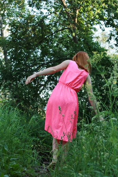 Una chica en un vestido rosa camina por el bosque, las manos tocan la hierba verde, la cara no es visible —  Fotos de Stock