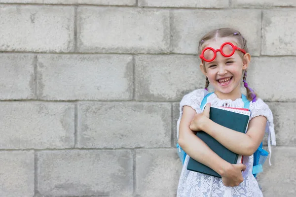 Glimlachend klein meisje met blond haar in rood speelgoed bril staat met boeken in haar handen en een rugzak op haar rug, schoolleven concept — Stockfoto