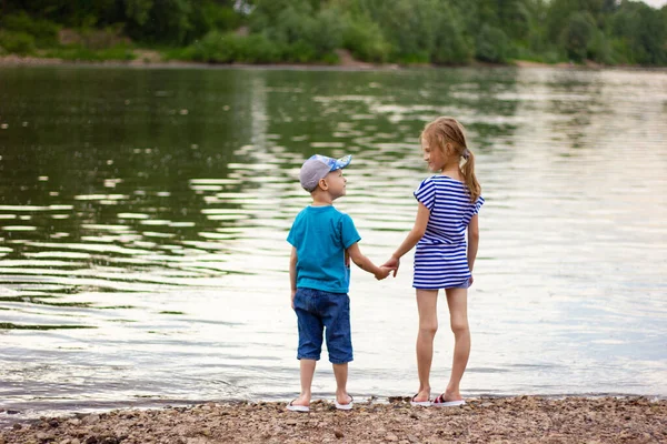 Брат и сестра держатся за руки и стоят на берегу реки летним вечером, детская дружба — стоковое фото
