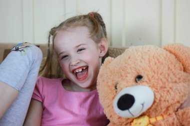 Pembe elbiseli, at kuyruklu, oyuncak ayıya sarılan, süt dişsiz gülümseyen küçük bir kızın portresi.