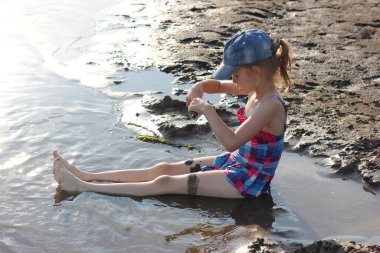 Parlak mayo giymiş mutlu bir kız nehirde oturur ve kumla oynar, yazın çocuklarla aktif tatiller yapar.