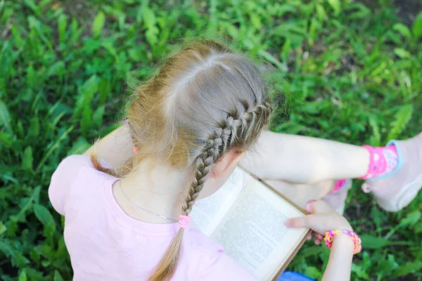 Klein blond meisje met vlechten in de zomer buiten in het groen leest een boek, foto van achteren — Stockfoto