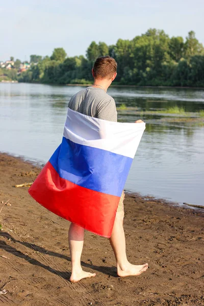 Un hombre está parado en la orilla del río con la bandera de Rusia de tres rayas blanco azul y rojo, una gran bandera rusa sobre los hombros de un hombre, el espíritu de libertad, patriotismo —  Fotos de Stock