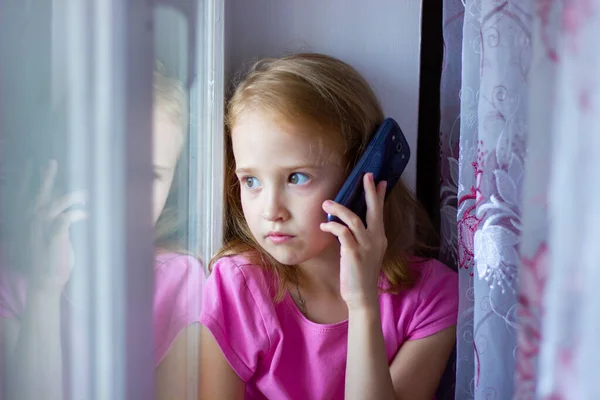 Pembe elbiseli küçük kız pencerenin önünde otururken telefonla konuşuyor, yeni teknolojiler.