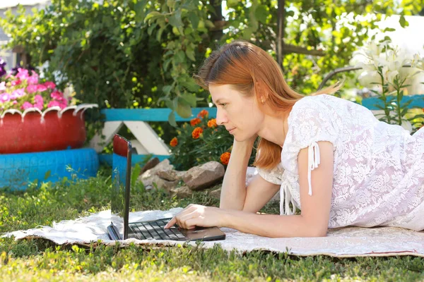 Kızıl saçlı bir kız diz üstü bilgisayarla kır evindeki çiçeklerin yanında yatıyor. Serbest çalışma, uzaktan eğitim, online alışveriş.