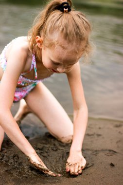 Kız nehir kıyısında elleri ile kum kazıyor, çocuklarla temiz havada yürüyüş yapıyor.