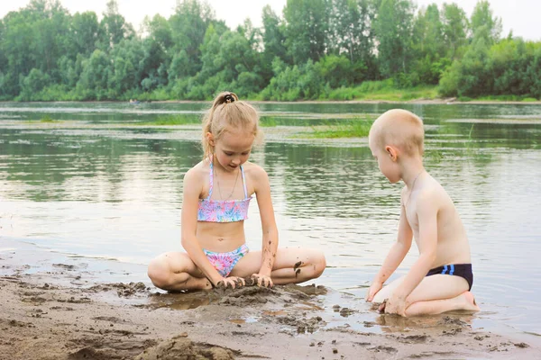Девочка и мальчик играют на песке у реки, брат и сестра отдыхают на природе — стоковое фото