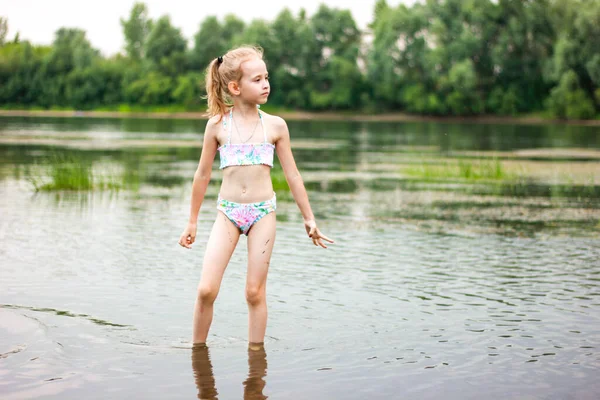 En liten flicka i baddräkt står i floden och tittar in i fjärran, sommarsemester med familj Royaltyfria Stockbilder
