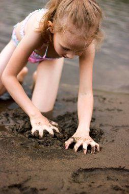 Kız nehir kıyısında elleri ile kum kazıyor, çocuklarla temiz havada yürüyüş yapıyor.