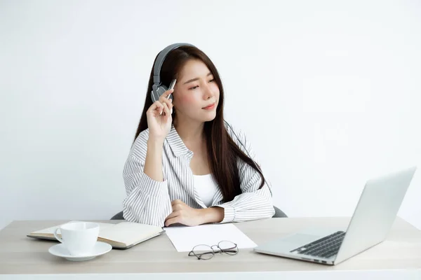 一位戴着耳机的年轻亚洲女性通过网络视频长途电话与一位朋友愉快地打招呼 她看着数字笔记本电脑屏幕 — 图库照片