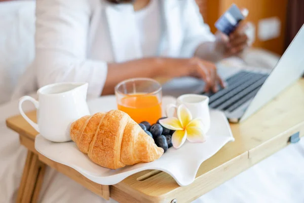 Focus on sweets fruit. A businesswoman was holding a credit card and using a laptop for online shopping and internet payments while relaxing in bed with snacks and fruit in the hotel room.