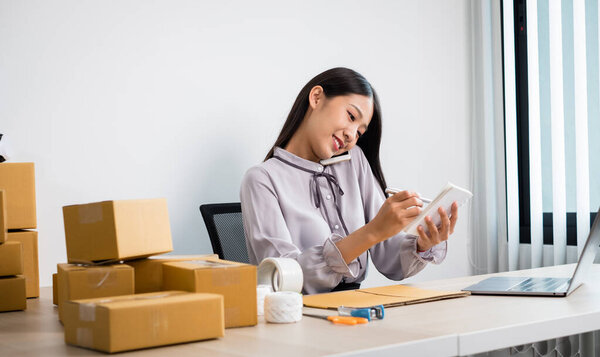 Young Asian woman business owner with many parcel boxes on the table happy online sales job, use your smartphone, get an order from customers, take notes, and make arrangements for delivery by post.