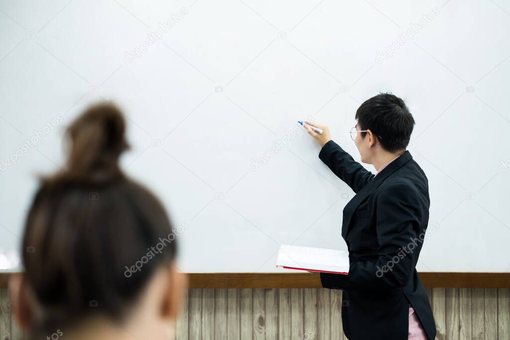 An Asian male teacher in a classroom near the whiteboard is writing and teaching a lesson with a special lecture in a class column. Math teacher explains arithmetic sums to university students.