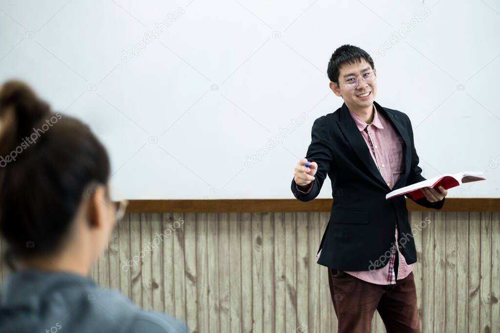 An Asian male teacher in a classroom near the whiteboard is writing and teaching a lesson with a special lecture in a class column. Math teacher explains arithmetic sums to university students.
