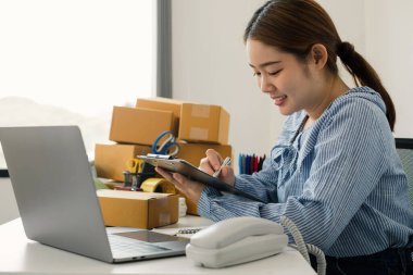 Genç Asyalı kadın işyeri sahibi masanın üzerinde mutlu online satış işi, dizüstü bilgisayarı kullan, müşterilerden emir al, notlar al ve teslimat için postayla ayarlamaları yap..