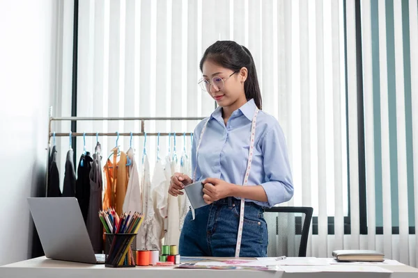 Tekstil aksesuarları ve girişimci tasarımcı çizimleriyle genç ve güzel Asyalı moda terzisi stüdyoda yaratıcı fikirler için dizüstü bilgisayarı olan parlak renklerle dolu..