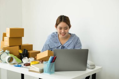 Genç Asyalı kadın işyeri sahibi masanın üzerinde mutlu online satış işi, dizüstü bilgisayarı kullan, müşterilerden emir al, notlar al ve teslimat için postayla ayarlamaları yap..